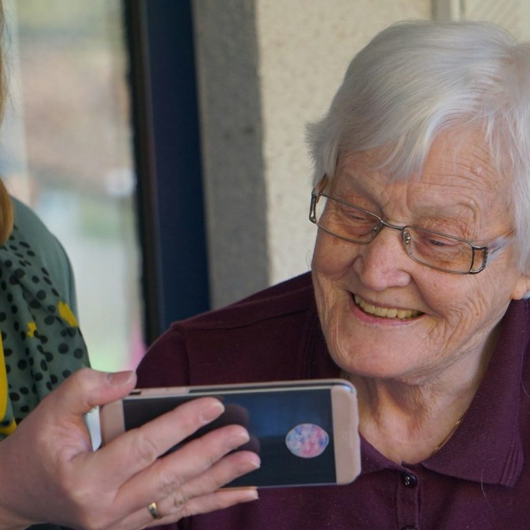 Teilhabe Hochaltrig Frau schaut auf ein Smartphone, welches sie von einer jüngeren Frau hingehalten bekommt.